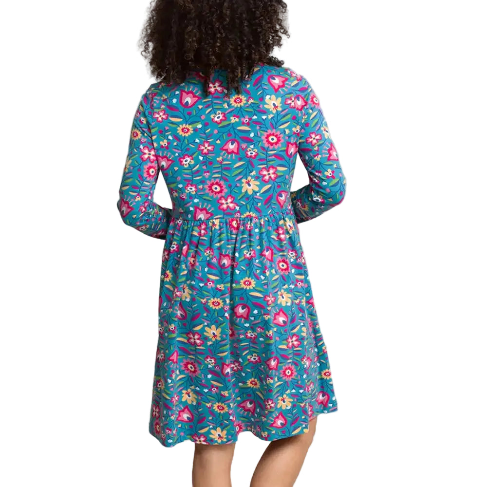 View of a woman shown from the back wearing a long-sleeve knee-length dress with a colorful floral print on a teal background, photographed against a plain light gray studio backdrop.
