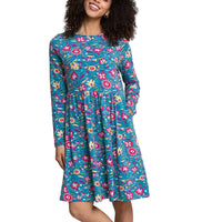 A woman wearing a long-sleeve, knee-length dress with a colorful floral print on a teal background, standing against a plain light gray studio backdrop.