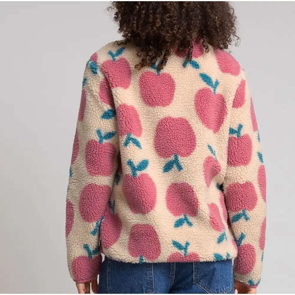 A person shown from the back wearing a cream sherpa fleece jacket with a pink apple pattern and blue leaf accents, paired with blue jeans, against a light gray studio background.