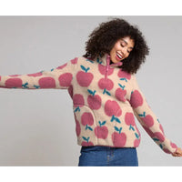A woman wearing a cream-colored fleece pullover sweater with a pink and teal apple pattern, paired with blue jeans, against a solid gray studio background.