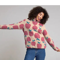 A woman wearing a cream-colored fleece pullover sweater with a pink and teal apple pattern, paired with blue jeans, posing against a solid gray studio background.