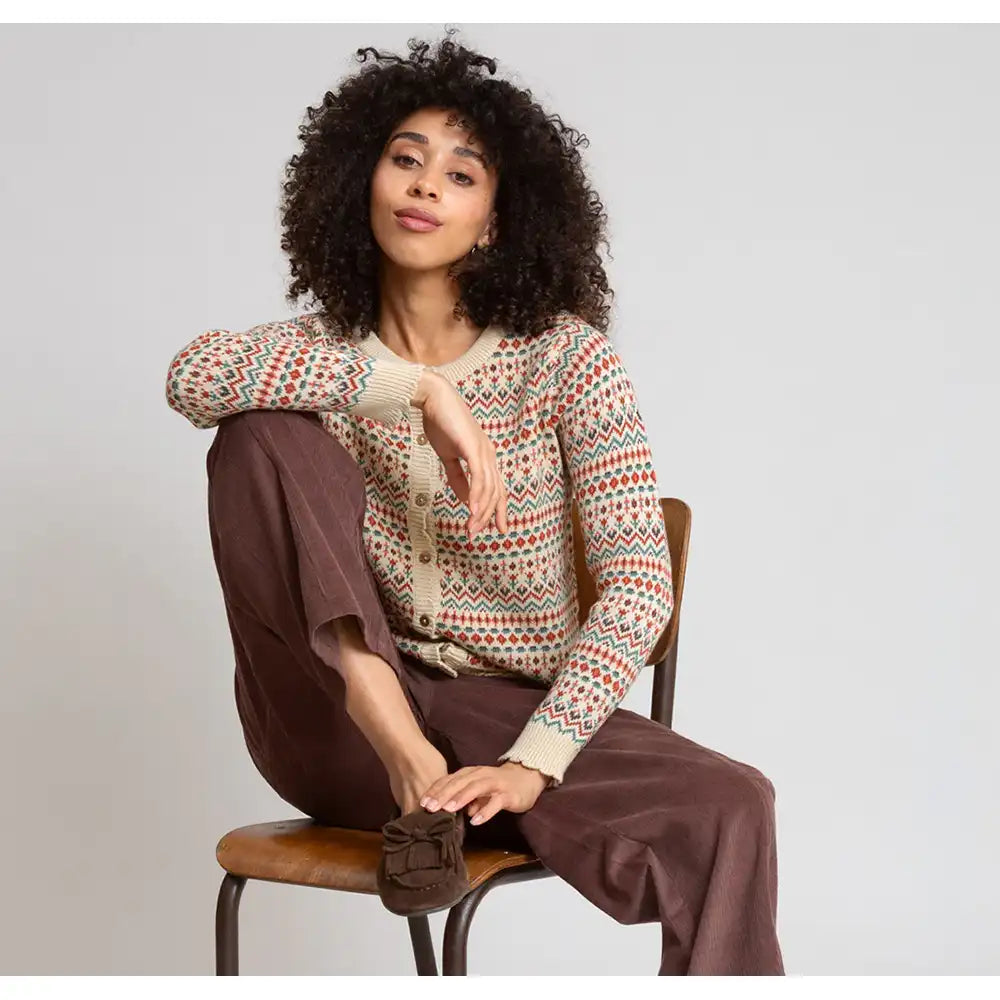 A woman with curly black hair sits on a wooden chair wearing a cream-colored knit cardigan with a multicolor geometric pattern, brown wide-leg trousers, and brown mule shoes against a light gray studio background.