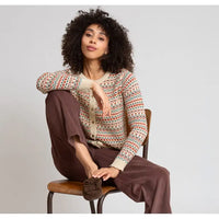 A woman with curly black hair sits on a wooden chair wearing a cream-colored knit cardigan with a multicolor geometric pattern, brown wide-leg trousers, and brown mule shoes against a light gray studio background.