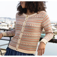 A woman wearing a cream-colored knit cardigan with a multicolor Fair Isle pattern of red, green, and blue geometric motifs, standing by a harbor railing.
