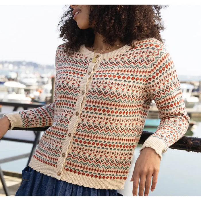 A woman wearing a cream-colored knit cardigan with a multicolor Fair Isle pattern of red, green, and blue geometric motifs, standing by a harbor railing.