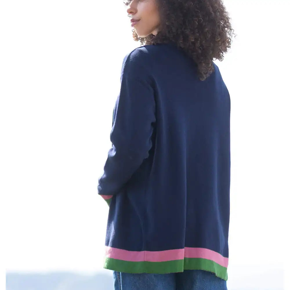 A woman shown from the back wearing a navy blue knit cardigan with pink and green striped trim at the hem, paired with blue jeans against a bright white outdoor background.