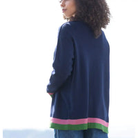 A woman shown from the back wearing a navy blue knit cardigan with pink and green striped trim at the hem, paired with blue jeans against a bright white outdoor background.