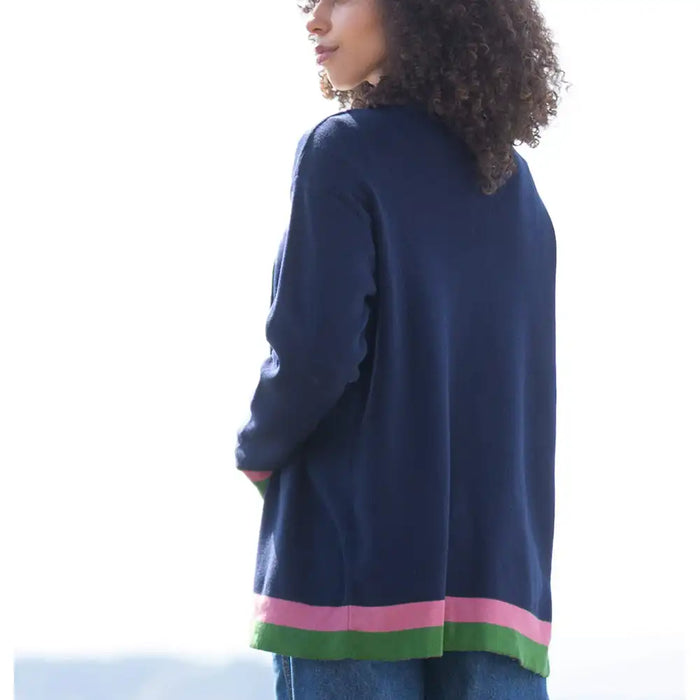 A woman shown from the back wearing a navy blue knit cardigan with pink and green striped trim at the hem, paired with blue jeans against a bright white outdoor background.