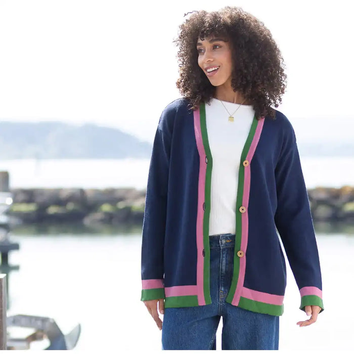 A woman wearing a navy blue cardigan with pink and green striped trim over a white top and blue jeans, standing outdoors near a waterfront with a blurred background.