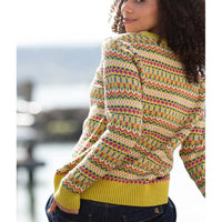 A woman wearing a colorful patterned knit sweater with yellow ribbed cuffs and hem, paired with dark blue jeans, photographed outdoors with a blurred waterfront background.