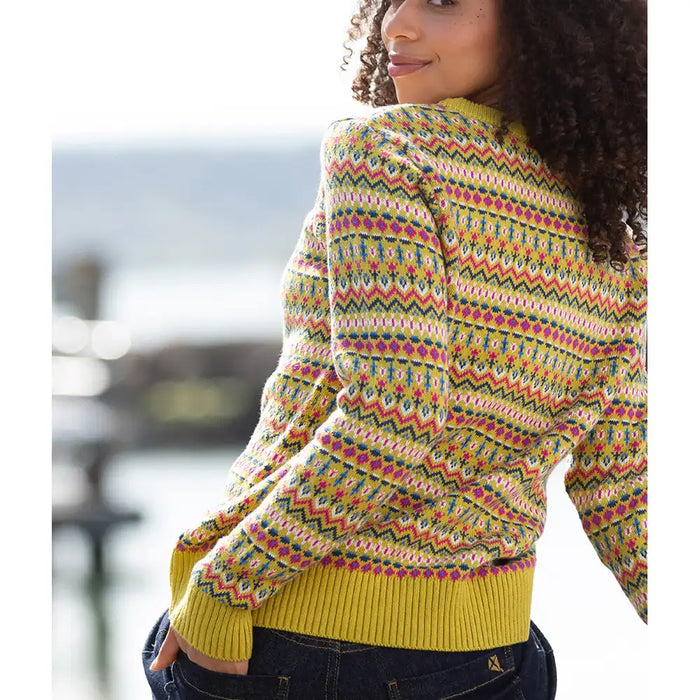 A woman wearing a colorful patterned knit sweater with yellow ribbed cuffs and hem, paired with dark blue jeans, photographed outdoors with a blurred waterfront background.