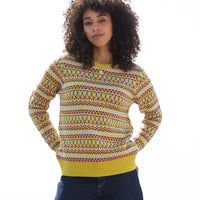 A woman with curly dark hair wearing a yellow and multicolor geometric patterned knit sweater, paired with dark blue jeans, against a plain white background.