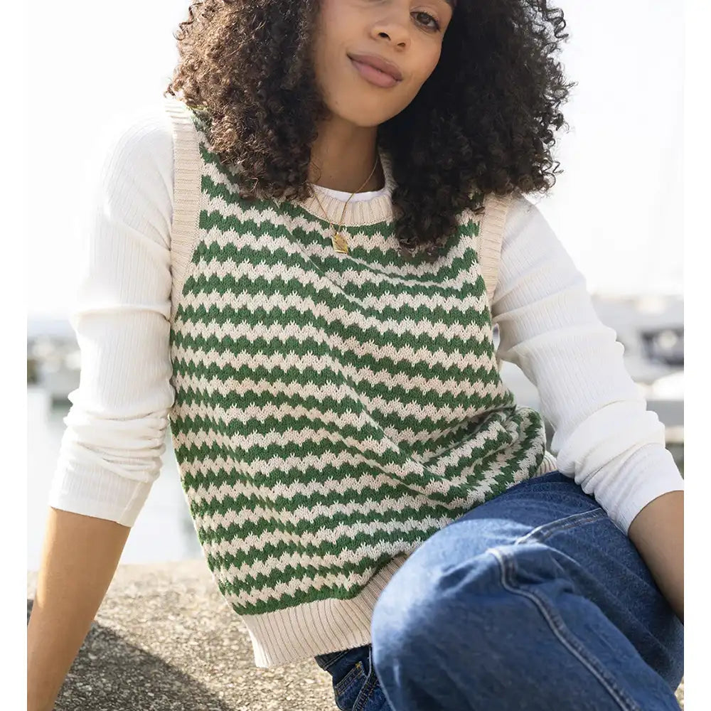 A woman wearing a green and cream zigzag knit sweater vest over a white long-sleeve shirt and blue jeans, sitting outdoors.