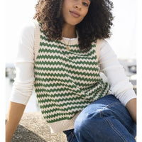 A woman wearing a green and cream zigzag knit sweater vest over a white long-sleeve shirt and blue jeans, sitting outdoors.