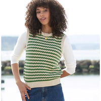 A woman wearing a green and cream chevron-patterned knit sweater vest over a cream long-sleeve shirt and blue jeans, standing outdoors with a blurred waterfront background.