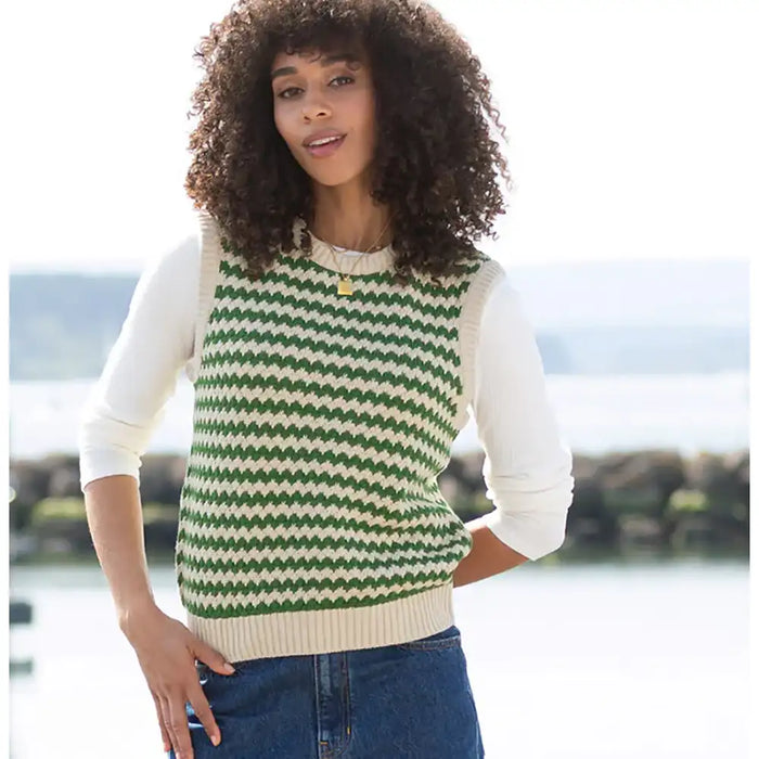 A woman wearing a green and cream chevron-patterned knit sweater vest over a cream long-sleeve shirt and blue jeans, standing outdoors with a blurred waterfront background.