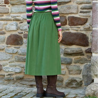 A back view of a woman wearing a green midi skirt, a striped long-sleeve top, and brown boots, standing in front of a rustic stone wall.