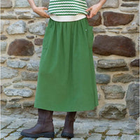 A person wearing a green cord midi skirt with side pockets, a green and white chevron sweater, and brown boots, standing in front of a rustic stone wall.