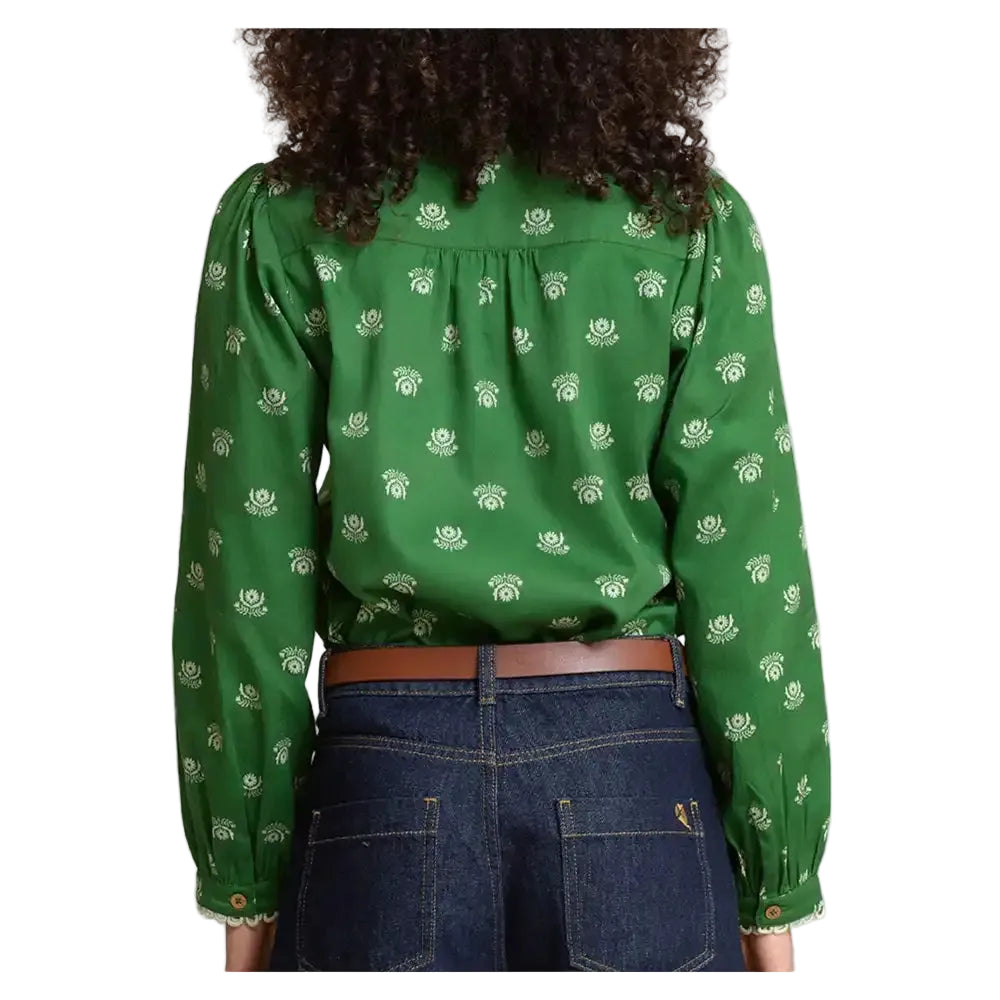 A woman shown from the back wearing a green long-sleeve blouse with a small white floral or leaf-like allover print, paired with dark blue jeans and a brown belt.
