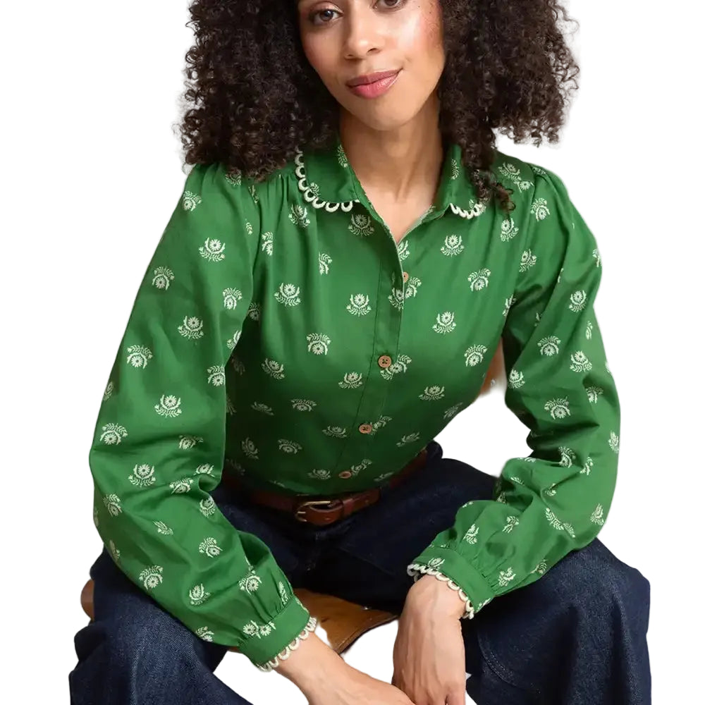 A woman wearing a green long-sleeve button-up blouse with a white floral print and scalloped collar, sitting on a wooden chair.