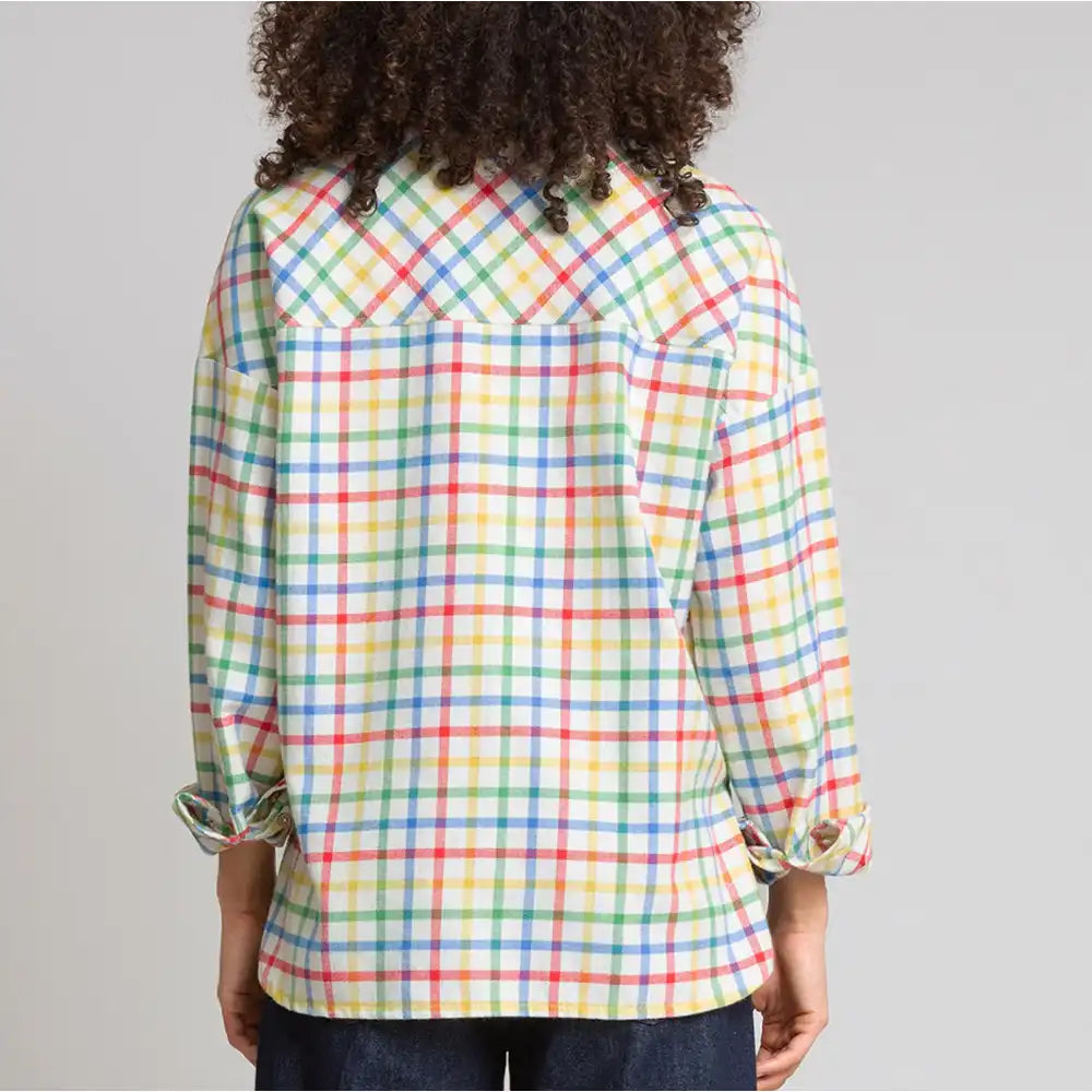 A person shown from the back wearing a long-sleeve plaid button-up shirt with a multicolor grid pattern on a light background, paired with dark jeans against a plain light gray studio backdrop.