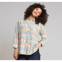 A woman with curly dark hair wearing a long-sleeve button-up plaid shirt in pastel rainbow colors, paired with dark jeans, posing against a solid gray studio background.