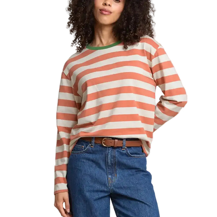 A woman wearing a long-sleeve crewneck top with horizontal coral and off-white stripes, paired with blue jeans and a brown belt, against a plain light gray studio background.