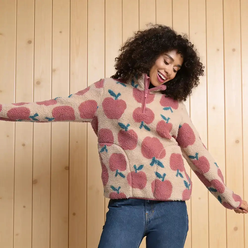 A woman wearing a cream sherpa fleece half-zip pullover sweater with a pink and blue cherry pattern, paired with blue jeans, posing against a light wood-paneled wall.