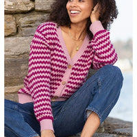 A woman wearing a pink and magenta chevron-patterned knit cardigan with brown buttons, paired with blue jeans, sitting outdoors against a stone wall.