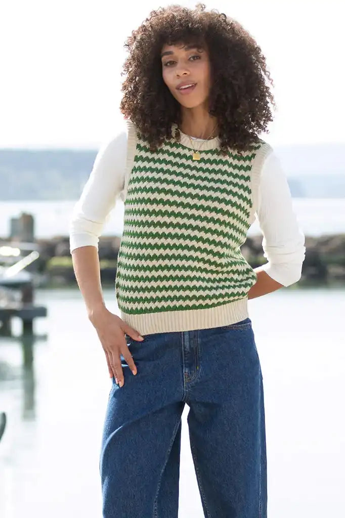 A woman with curly dark hair models a sleeveless knit sweater vest featuring a green and cream chevron pattern over a cream long-sleeve shirt, paired with blue jeans, set against a bright, blurred outdoor background.