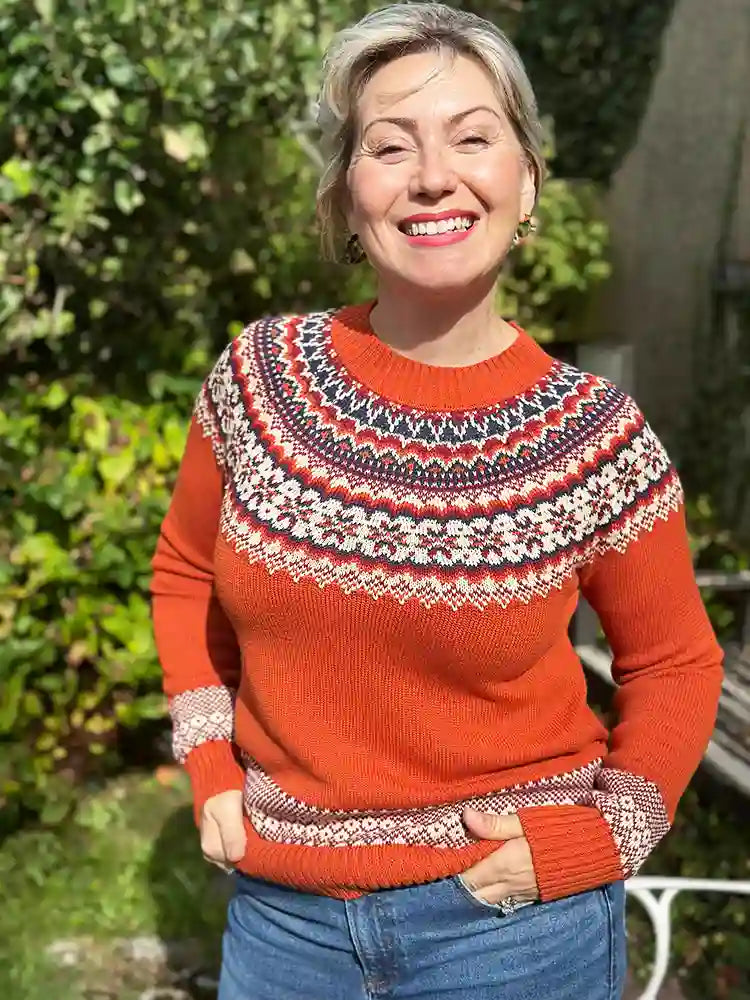 A smiling woman wearing a bright orange Fair Isle knit sweater with intricate red, navy, and cream patterns across the yoke, cuffs, and hem, paired with blue jeans in an outdoor garden setting.