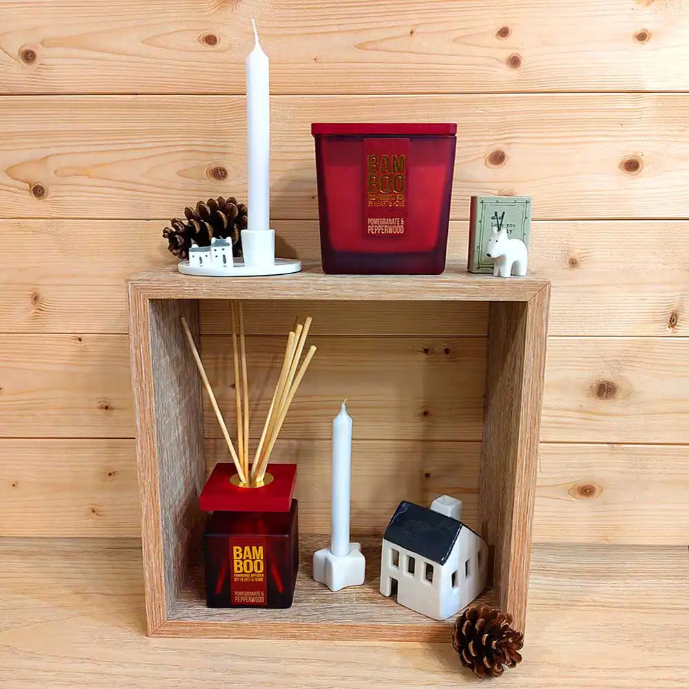 A styled holiday home decor display featuring a red scented candle, a bamboo reed diffuser, a white taper candle, and miniature white house figurines, all arranged on a light wood shelf against a wood-paneled wall.