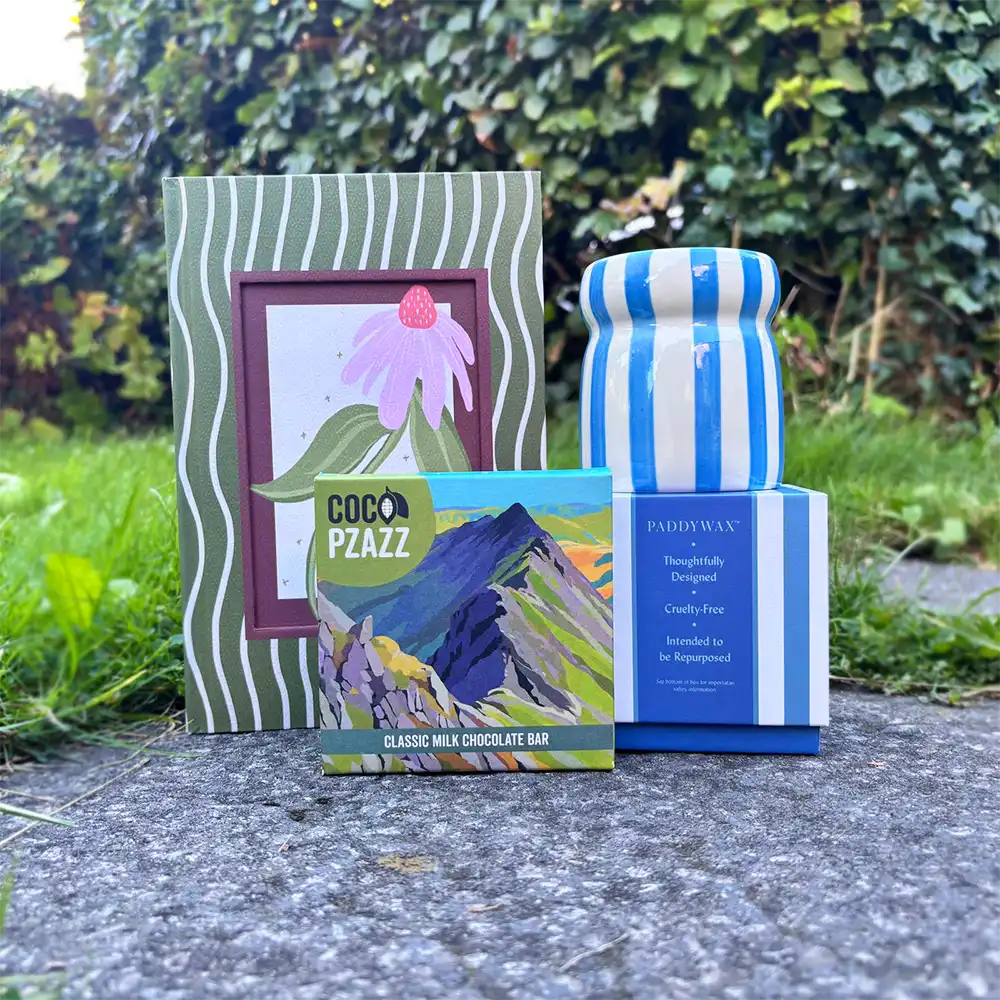 Lifestyle and giftware displayed outdoors on a stone surface with green foliage in the background: a square box of Coco PzaZZ chocolate bar, a blue-and-white striped box with a curve canfle on top and a green wavy stripped notebook witha magnolia flower in the middle. 