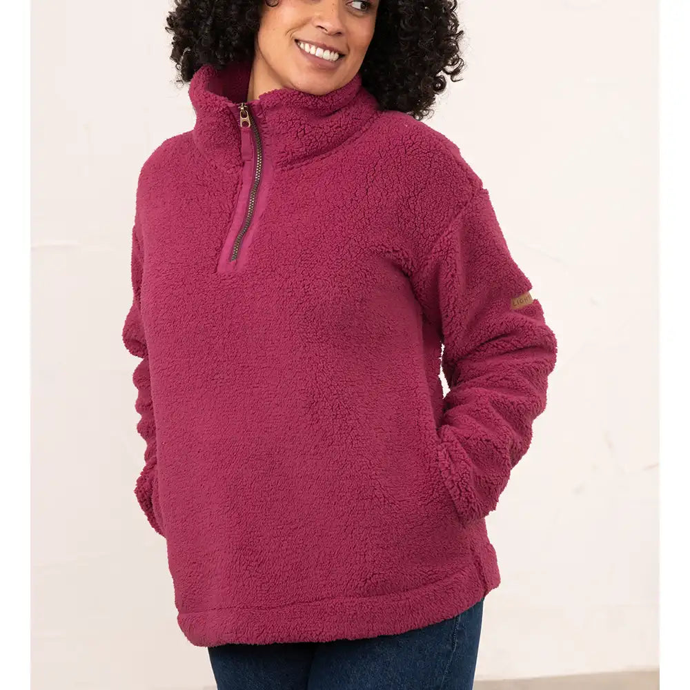 A woman wearing a dusty rose pink sherpa fleece quarter-zip pullover sweater with a mock neck and side pockets, paired with dark blue jeans, against a plain off-white wall.