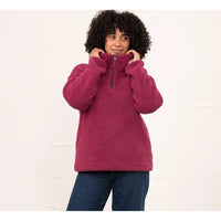 A woman with curly black hair wearing a dusty rose fleece quarter-zip pullover sweater and dark blue jeans, standing against a white textured wall.