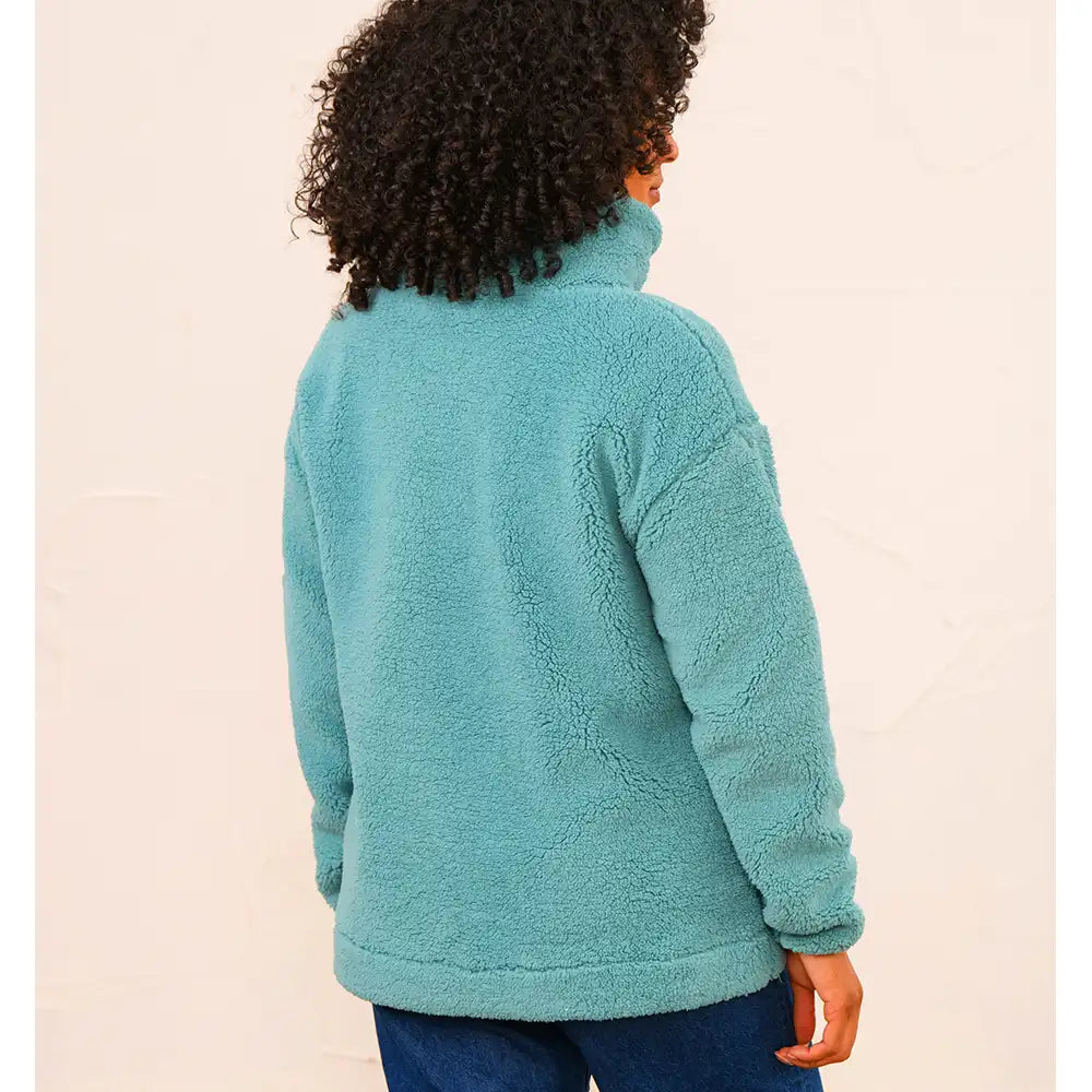 A person shown from the back wearing a plush, high-collar fleece pullover in a muted teal blue color, paired with dark blue jeans, against a plain light beige background.