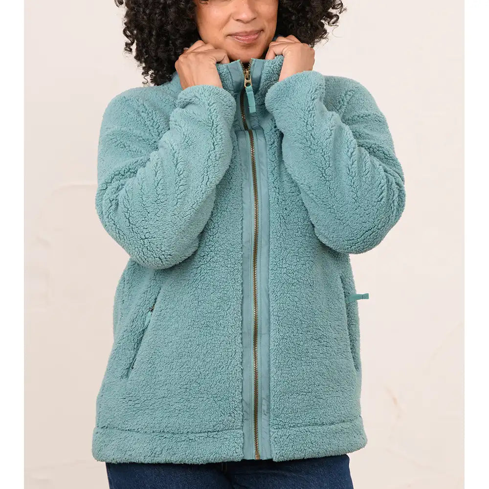 A woman wearing a teal sherpa fleece zip-up jacket, holding the collar closed. The jacket features a textured cable-knit pattern on the sleeves, a full front zipper, and side pockets, shown against a plain light beige background.