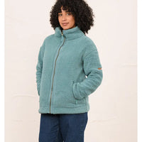 A woman wearing a teal sherpa fleece zip-up jacket with a high collar and side pockets, paired with dark blue jeans, standing against a light beige textured wall.