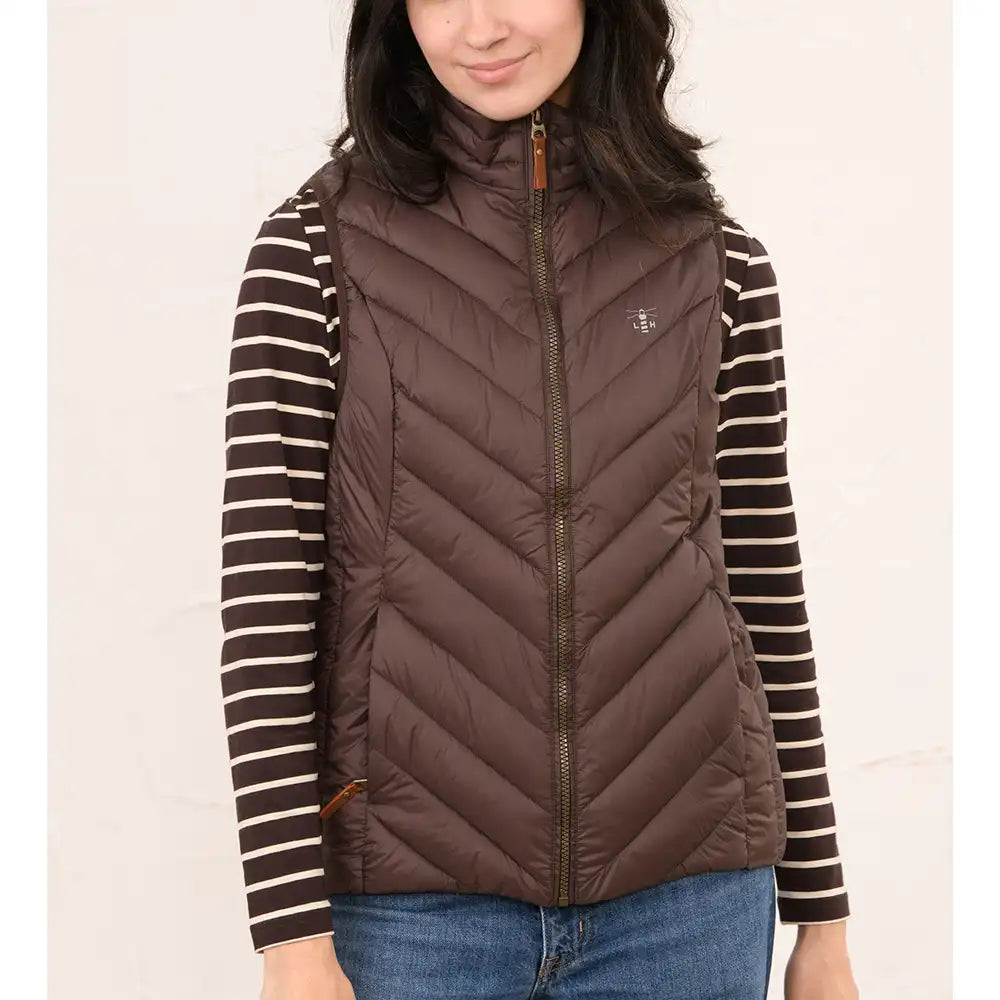 A woman wearing a brown puffer vest over a brown and cream striped long-sleeve shirt, paired with blue jeans, against a plain light background.