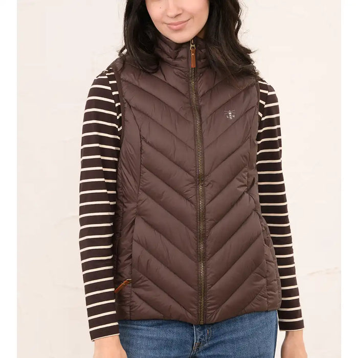A woman wearing a brown puffer vest over a brown and cream striped long-sleeve shirt, paired with blue jeans, against a plain light background.