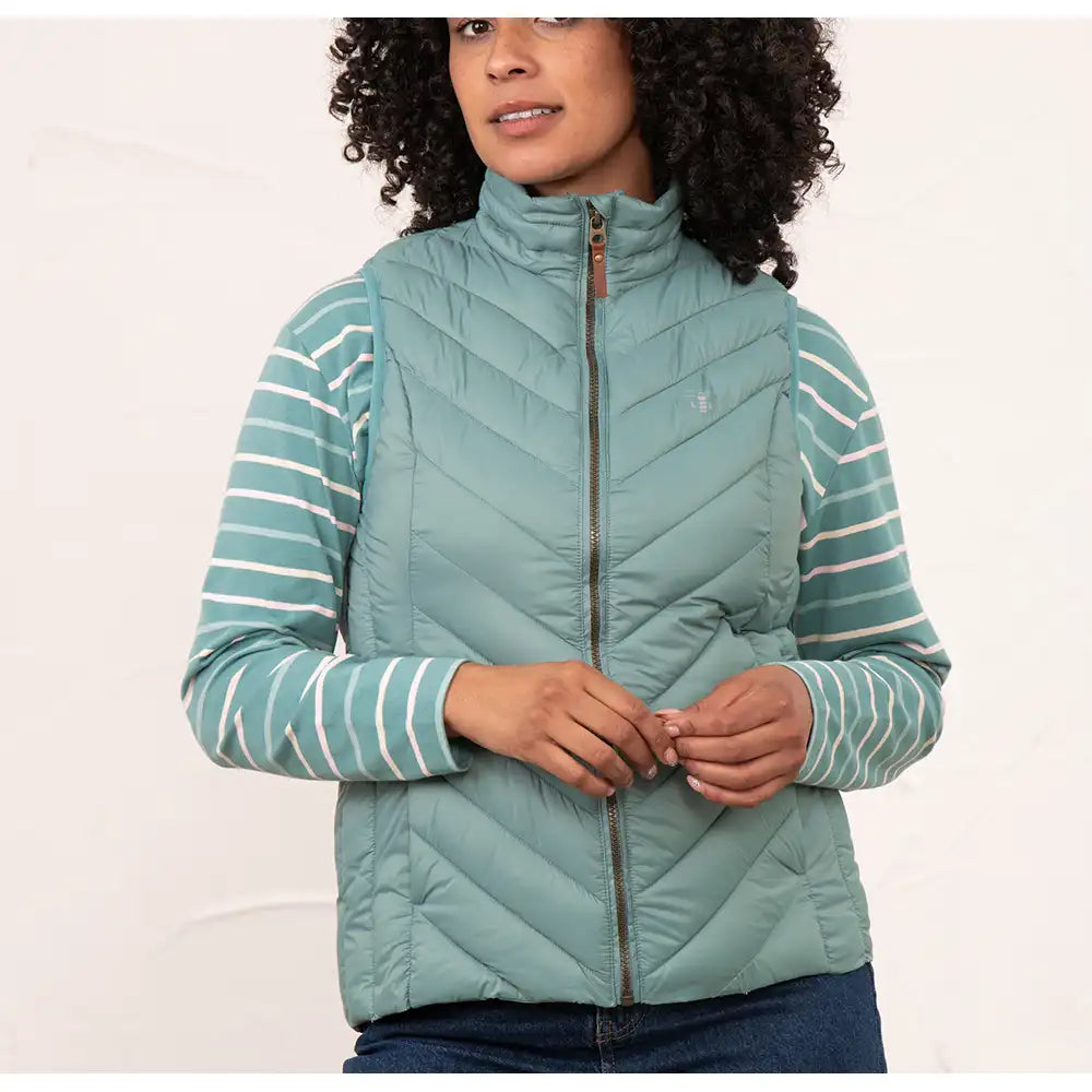 A woman wearing a sage green quilted puffer vest over a matching sage green and white striped long-sleeve top, paired with dark blue jeans, against a plain white studio background.