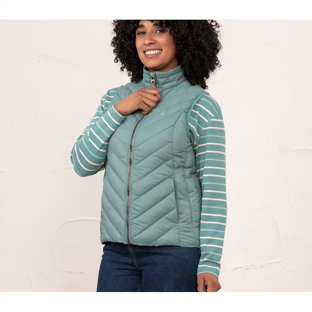 A woman wearing a sage green quilted zip-up puffer vest over a matching sage green and white striped long-sleeve shirt, paired with dark blue jeans, standing against a textured off-white wall.