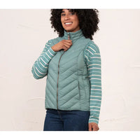 A woman wearing a sage green quilted zip-up puffer vest over a matching sage green and white striped long-sleeve shirt, paired with dark blue jeans, standing against a textured off-white wall.