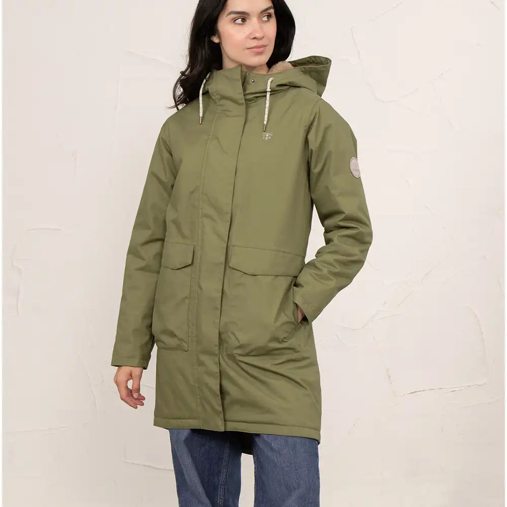 A woman wearing a mid-length olive green hooded parka jacket with front flap pockets and drawstrings, paired with blue jeans, standing against a textured off-white wall.