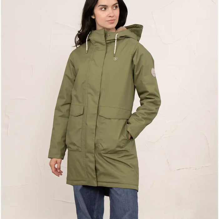 A woman wearing a mid-length olive green hooded parka jacket with front flap pockets and drawstrings, paired with blue jeans, standing against a textured off-white wall.
