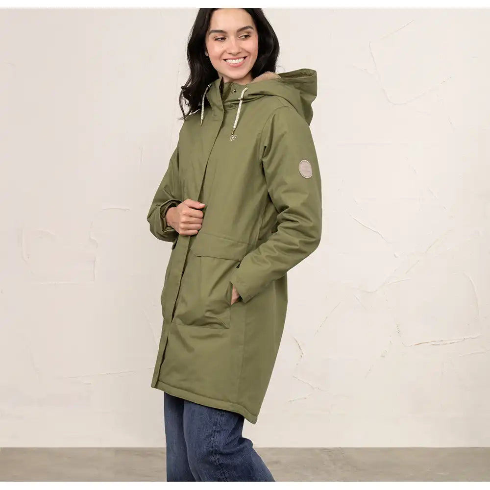 A woman wearing a mid-length olive green hooded parka jacket with front pockets, paired with blue jeans, standing against a textured off-white wall.