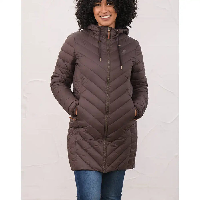A woman wearing a brown hooded quilted puffer jacket with a front zipper and drawstrings, paired with blue jeans, standing against a light textured wall.