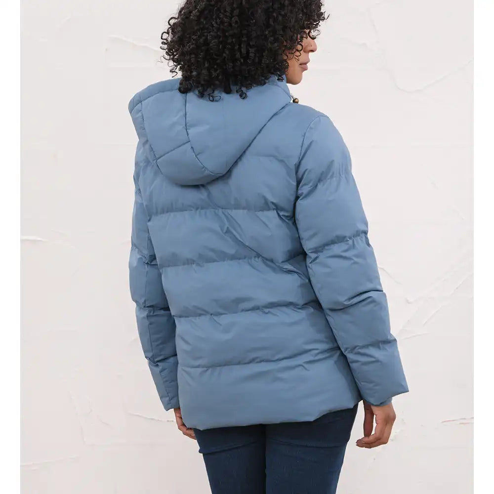 A woman wearing a dusty blue puffer jacket with a hood, shown from the back against a textured off-white wall.