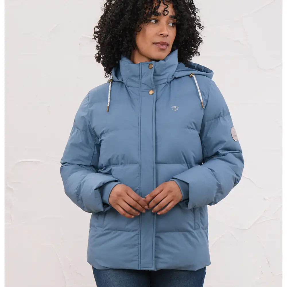 A woman wearing a light blue hooded puffer jacket with a high collar, front zipper, and drawstrings, standing against a textured off-white wall.