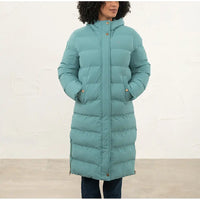 A woman wearing a long, hooded puffer coat in a muted teal-green color, standing against a light off-white wall.